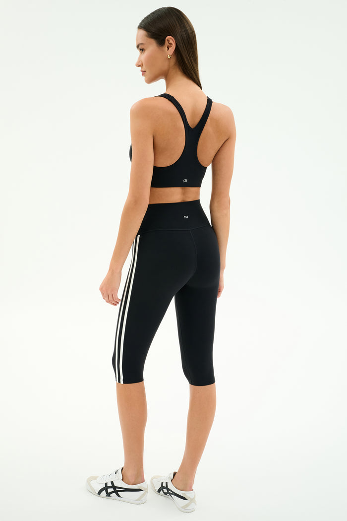 A woman stands in a three-quarter pose wearing the SPLITS59 Ella High Waist Airweight Capri in Black/White and a black sports bra with white sneakers against a plain background.