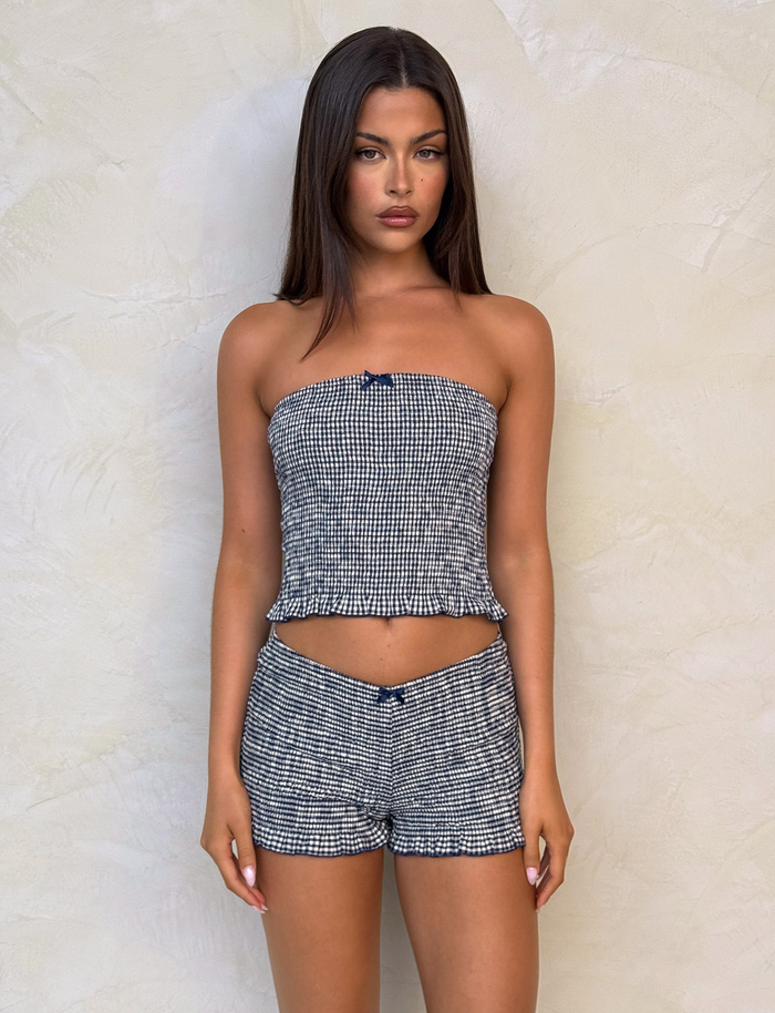 A woman with straight brown hair stands against a beige wall, wearing TIGERMIST's ERYNN SHORTS in navy gingham and a matching strapless top. She poses with a neutral expression and her arms by her sides.
