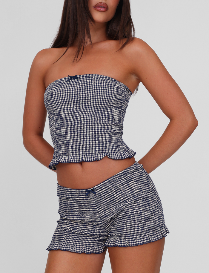 A woman wears the TIGERMIST ERYNN SHORTS in navy gingham with a matching strapless top, featuring small bow accents and a ruffled hem, set against a plain light background.