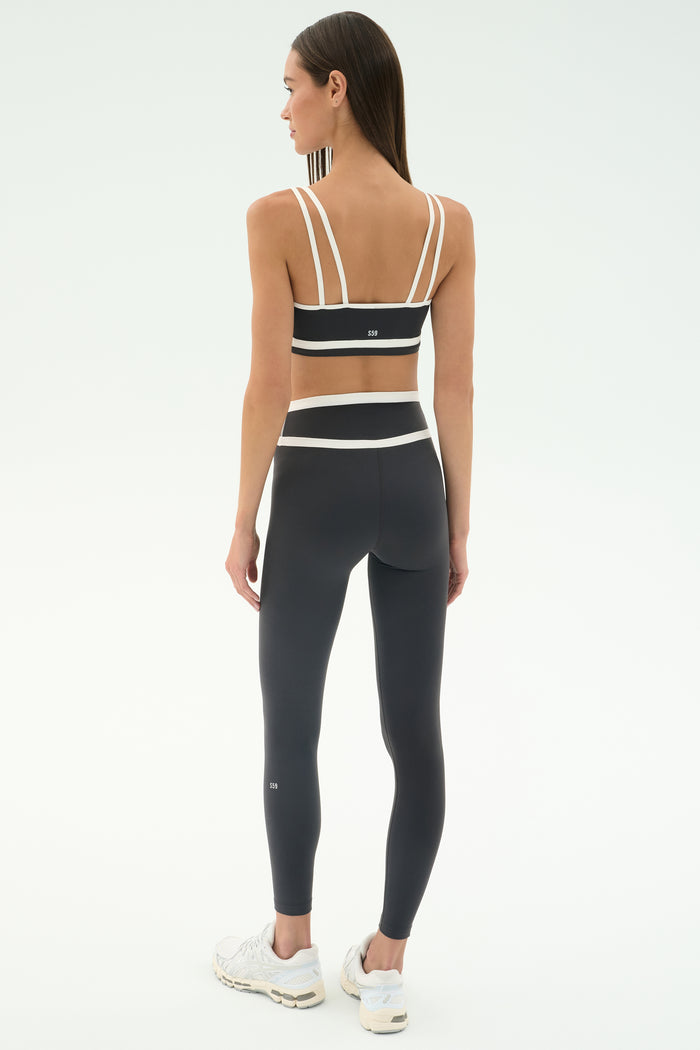 A woman faces away in the SPLITS59 Eva Rigor Double Strap Bra in Graphite/White, paired with high-waisted leggings and white sneakers—an ideal gym-ready look—against a plain white background.