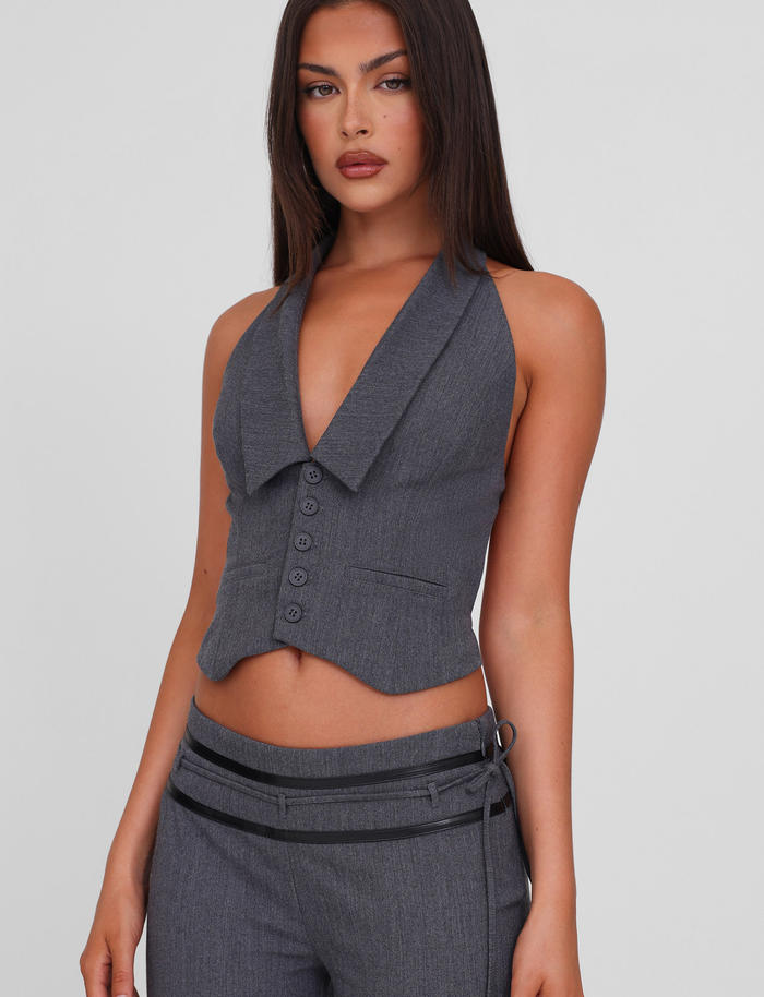A woman with long brown hair wears the TIGERMIST FEDRA VEST - GREY, a tailored, sleeveless vest with a deep V-neck and matching pants styled with a black belt and decorative straps, standing against a plain light background.