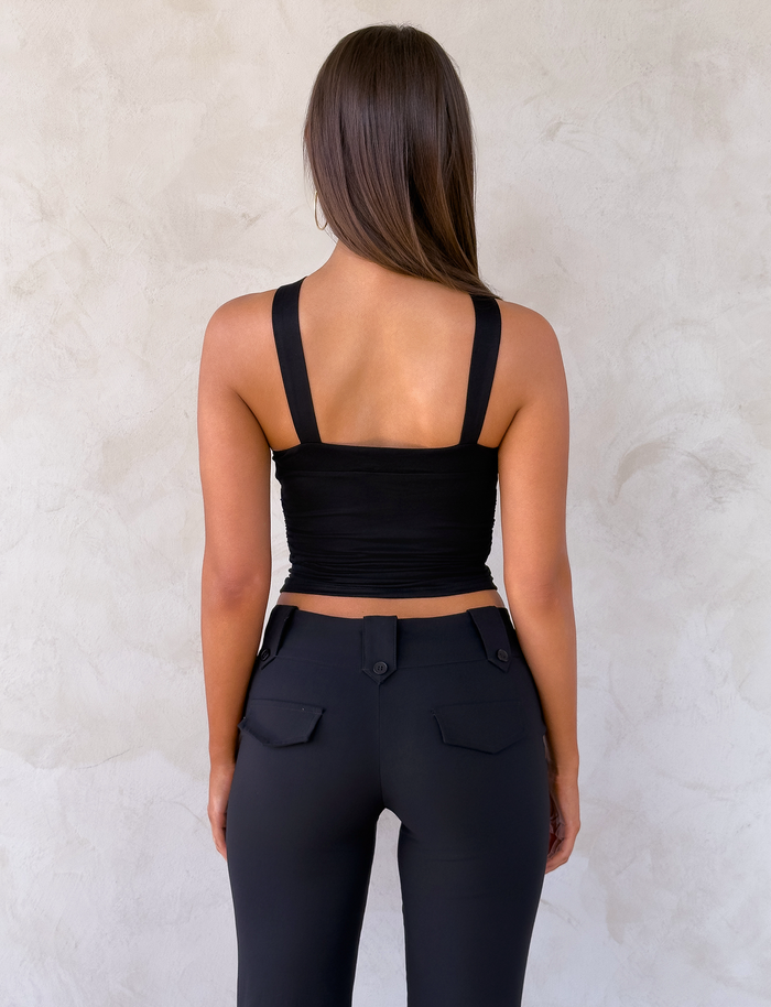 A woman with long brown hair stands facing a textured light wall, wearing TIGERMIST's FINCH TOP in black and black flap-pocket pants. Her arms are by her sides.
