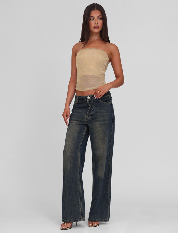 A woman with long straight hair and a neutral expression poses against a plain background, wearing the TIGERMIST HARRIETT TUBE TOP in gold, high-waisted wide-leg dark blue jeans, and open-toe heels.