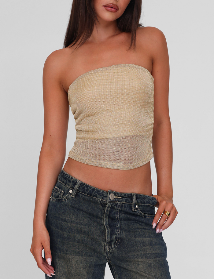 A woman wearing the TIGERMIST HARRIETT TUBE TOP in gold and low-rise blue jeans stands against a plain light background. Her left hand is on her hip, and she wears a gold ring on her right hand.