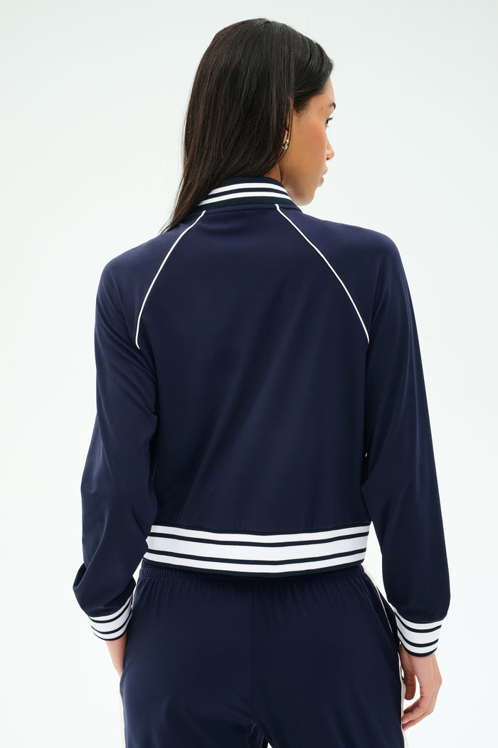 A person with long dark hair is shown from the back wearing the SPLITS59 Hayes Varsity Rigor Jacket in Indigo, made from stretch fabric, against a plain light background.