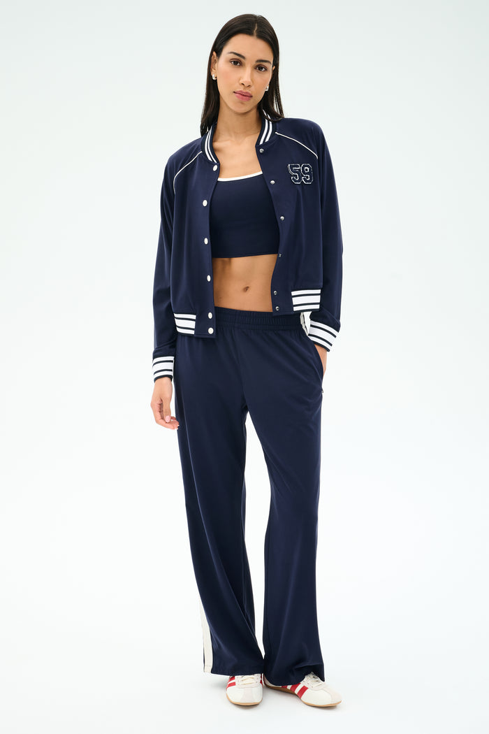A woman wears the SPLITS59 Hayes Varsity Rigor Jacket in Indigo, paired with a matching crop top and loose stretch pants with white stripes, and white sneakers, standing against a plain light background.
