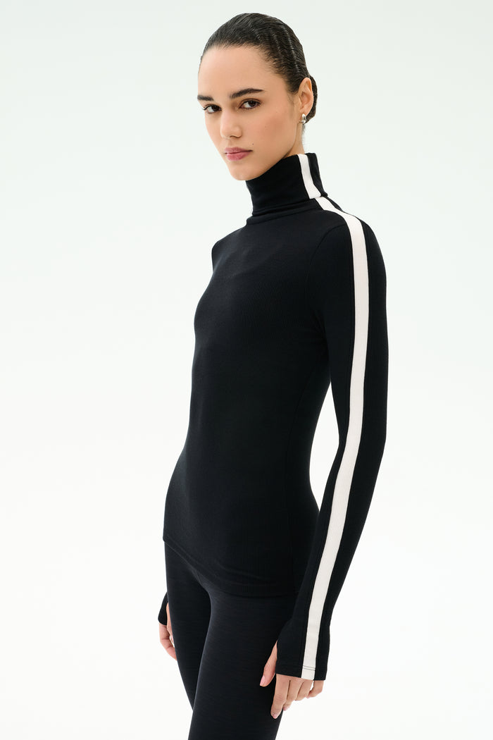 A woman stands against a plain light background wearing the SPLITS59 Jackson Rib Full Length Turtleneck in Black/White with a fitted design and white vertical sleeve stripe.