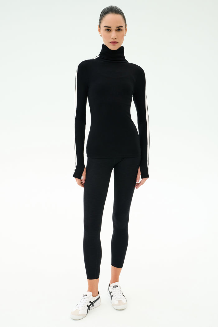 A woman faces forward in a SPLITS59 Jackson Rib Full Length Turtleneck - Black/White, paired with black leggings and white sneakers, set against a plain white background.