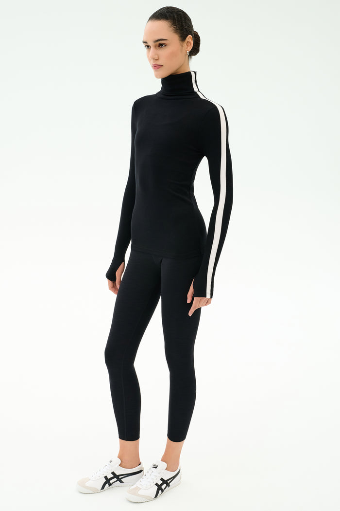 A person wears the SPLITS59 Jackson Rib Full Length Turtleneck in Black/White, paired with black leggings and white sneakers, standing against a plain white background.