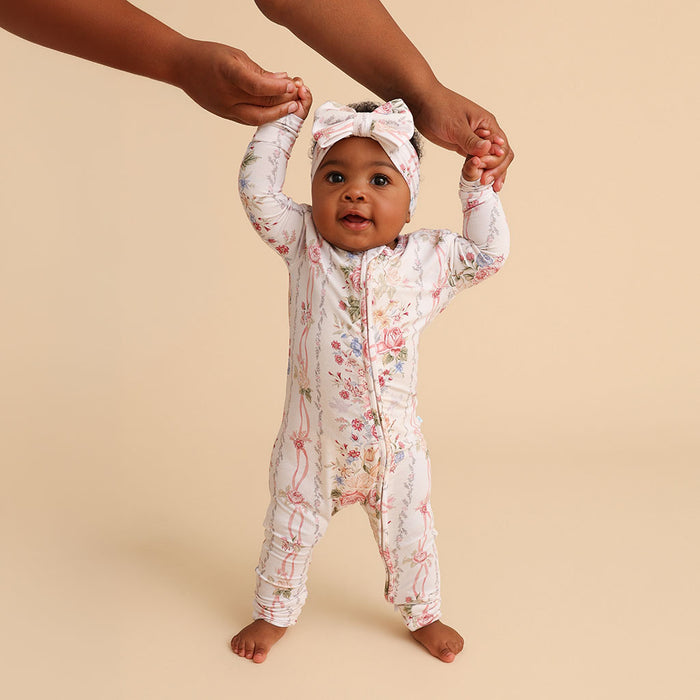 Baby standing, supported by an adult's hands, wearing the Janey print convertible one-piece