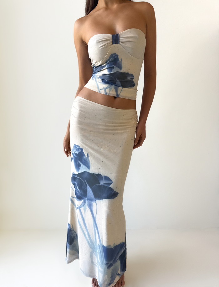 A woman models the TIGERMIST JANIS MAXI SKIRT in Indigo Floral, a beige skirt with a fitted waist and long silhouette featuring bold blue florals, standing against a plain white background.