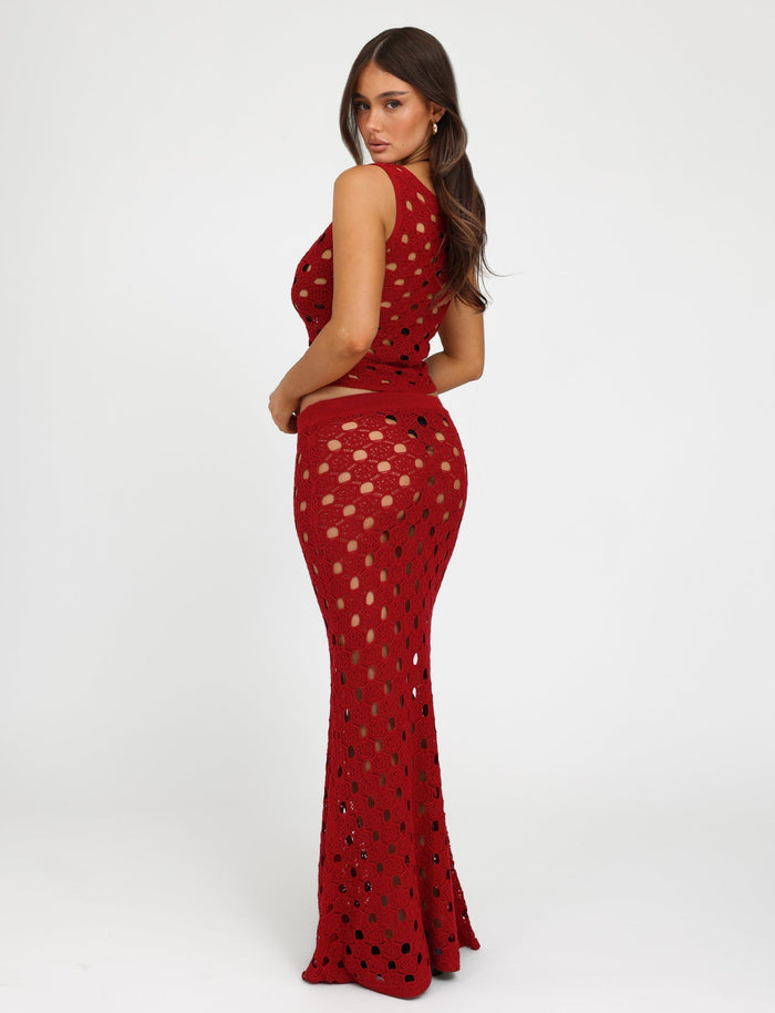 A woman with long brown hair is posing sideways and looking at the camera. She is wearing a cherry red, sleeveless, full-length crochet dress featuring circular patterns from Tiger Mist. The dress reveals a two-piece swimsuit underneath, against a plain white background.
