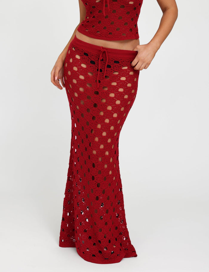 A person modeling the TIGERMIST KINCADE SKIRT in Cherry Red, featuring a red crochet maxi design with a drawstring waist. The fabric has an open, see-through pattern that reveals the legs beneath. The person is also partially showing a matching crochet top, set against a plain white background.