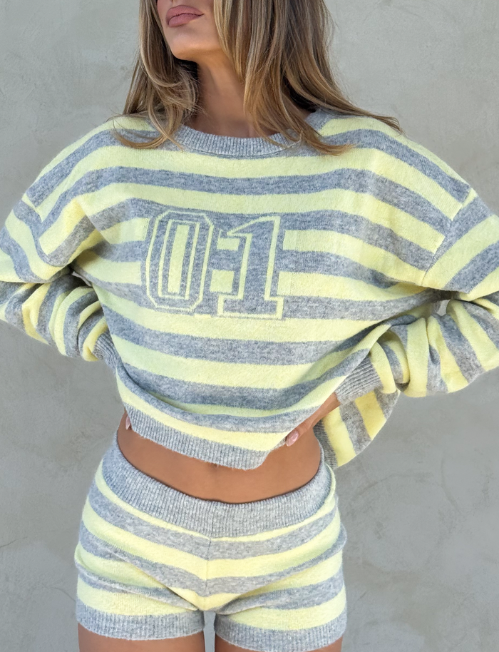A woman wears the TIGERMIST KOA JUMPER in yellow and grey stripes, featuring a bold "01" sporty print, paired with matching shorts, posing against a plain light-colored wall with her hands on her waist.