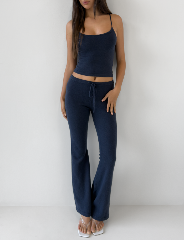 A woman poses against a white wall wearing TIGERMIST’s LEORA PANT in navy, featuring a relaxed fit and drawstring waist, styled with a navy crop top and white flip-flops. Her left hand is on her hip; her face is partially out of frame.