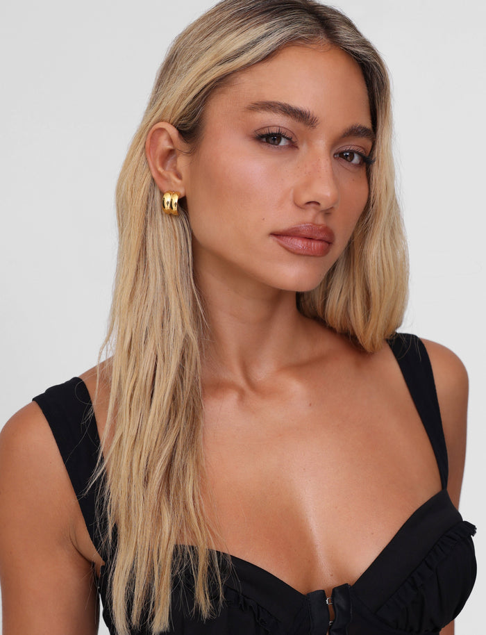A woman with long blonde hair, in a sleeveless black top, wears the stunning TIGERMIST LILLIA EARRING in 18k gold, looking directly at the camera against a plain background.