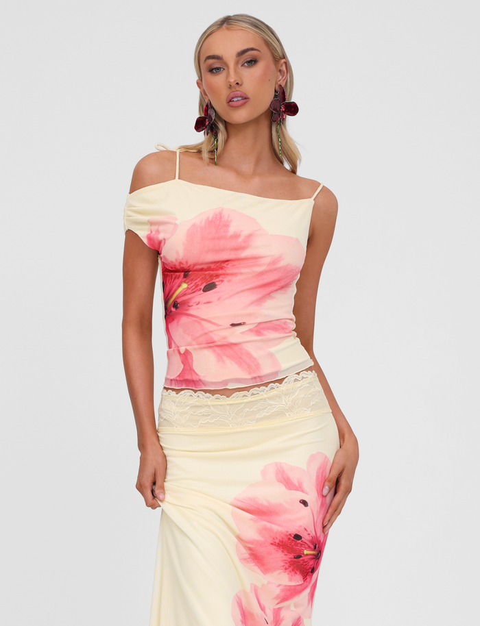 A woman poses in the TIGERMIST LILLIAN TOP - YELLOW, an off-shoulder style with bold floral design and lace waist trim, accessorized with statement red earrings against a plain light background.