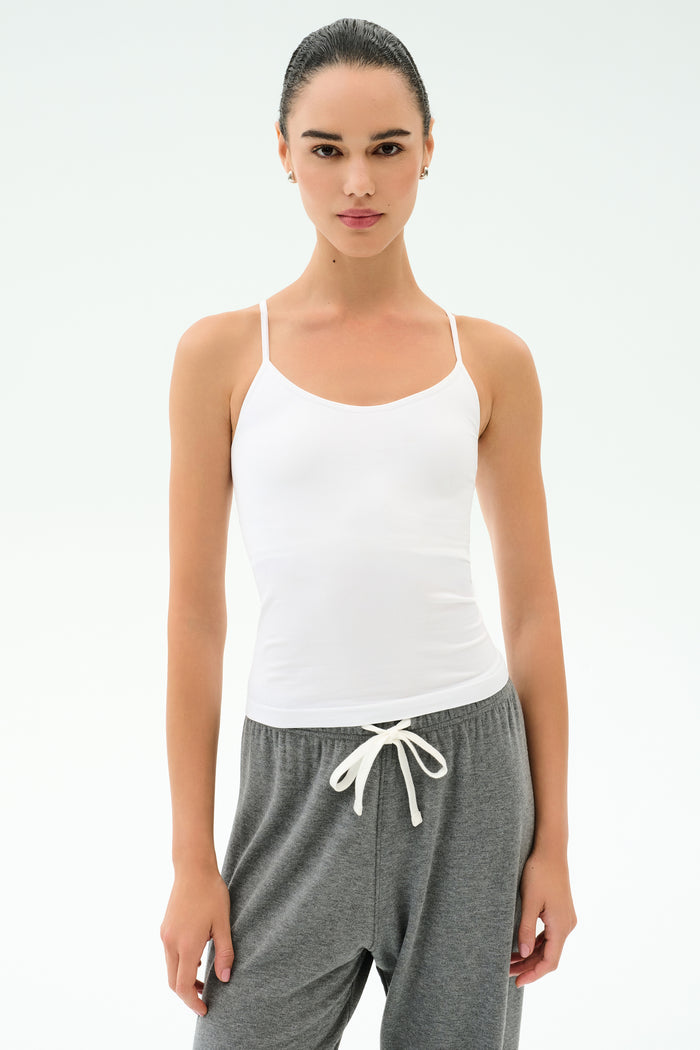 A woman wears the SPLITS59 Loren Seamless Waist Length Tank in white with gray sweatpants, standing facing forward against a plain white background.