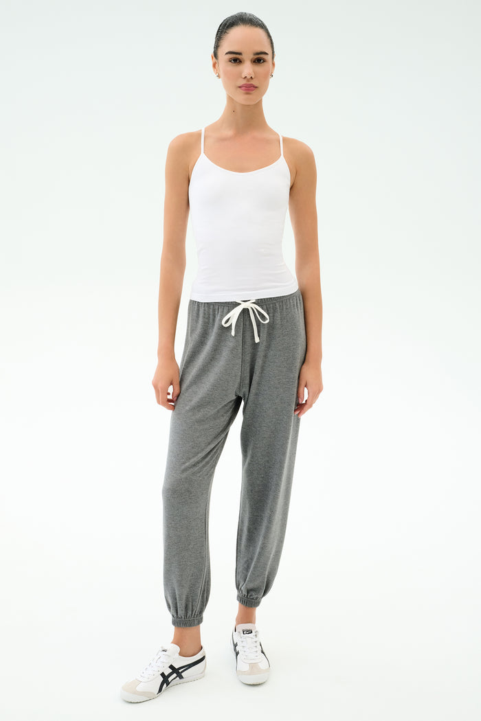 A woman faces forward on a white background, wearing the SPLITS59 Loren Seamless Waist Length Tank in white, paired with gray drawstring jogger pants and white sneakers.