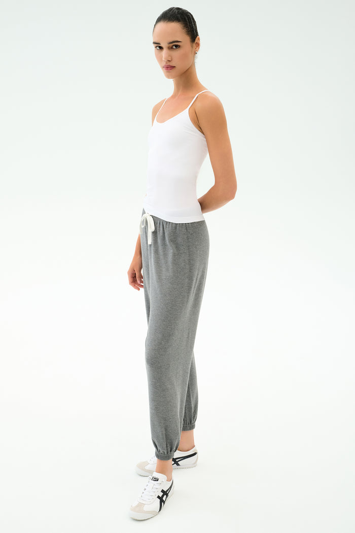Person standing against a plain background, wearing the SPLITS59 Loren Seamless Waist Length Tank in white, gray drawstring sweatpants, and white sneakers with black accents.