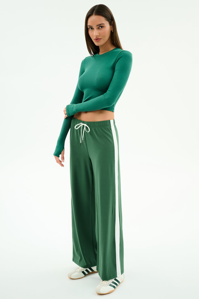 A woman wears the SPLITS59 Louise Rib Long Sleeve Crop in Arugula, paired with green wide-leg track pants featuring white side stripes and white sneakers with green accents, standing against a plain white background.