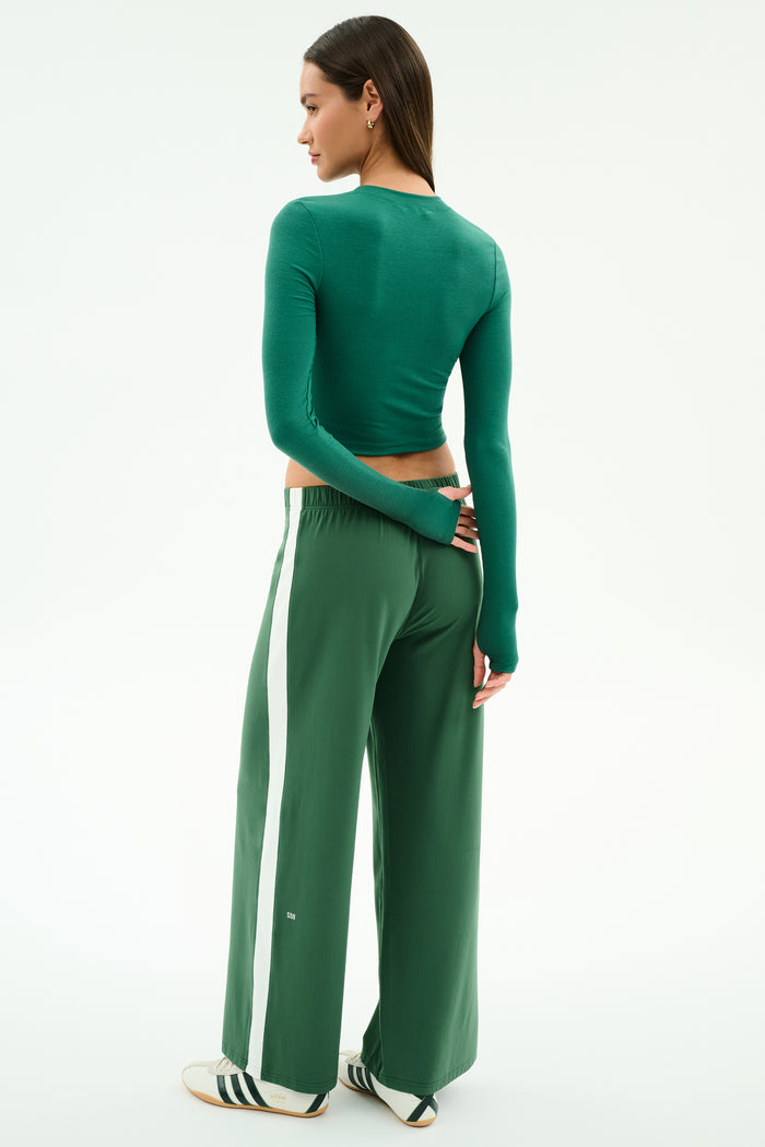 A woman stands facing away in the SPLITS59 Louise Rib Long Sleeve Crop in arugula and matching green wide-leg pants with a white stripe, paired with white sneakers featuring black stripes, set against a plain background.