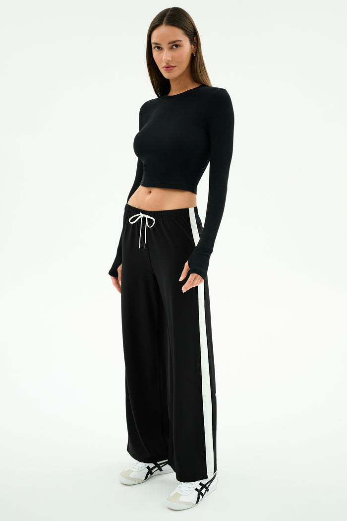 A woman stands facing slightly to the side, wearing the SPLITS59 Louise Rib Long Sleeve Crop in black, paired with black wide-leg pants featuring white side stripes and white sneakers, against a plain white background.