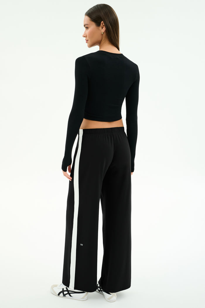 A woman stands facing away against a white background, wearing the SPLITS59 Louise Rib Long Sleeve Crop in black, paired with loose black pants with white side stripes and white sneakers.