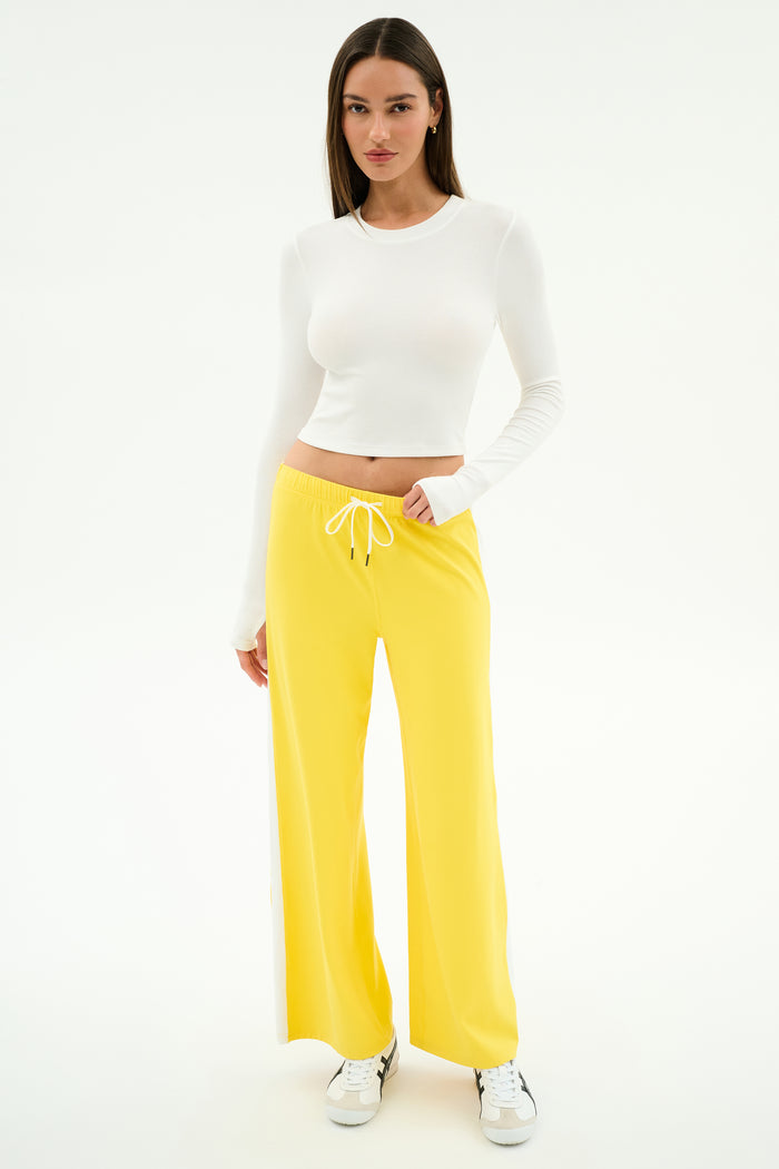 A woman stands in a studio wearing the SPLITS59 Louise Rib Long Sleeve Crop in white, paired with yellow wide-leg drawstring pants and white sneakers—an ideal gym-ready look—against a plain white background.