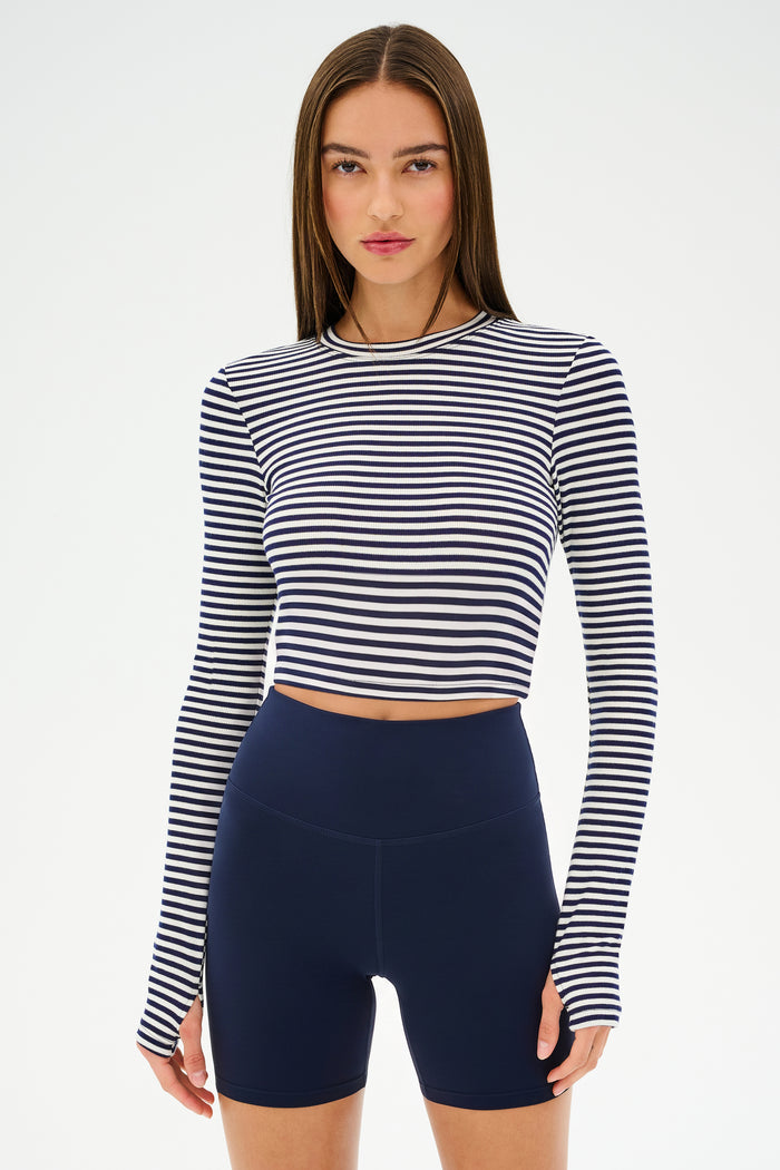 A woman wears the SPLITS59 Louise Rib Long Sleeve Crop in White/Indigo Stripe with high-waisted navy blue bike shorts, standing against a plain white background.