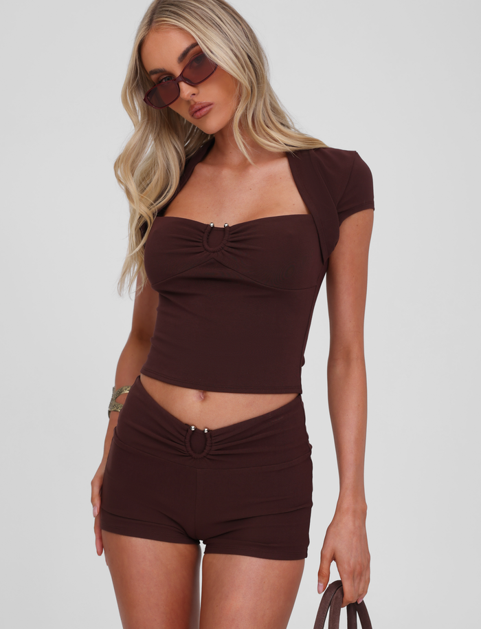 A woman with long blonde hair wears brown sunglasses and a matching cropped tee with the TIGERMIST LUNARA SHORT – CHOCOLATE, featuring a gathered waistband, holding a bag against a plain light background.