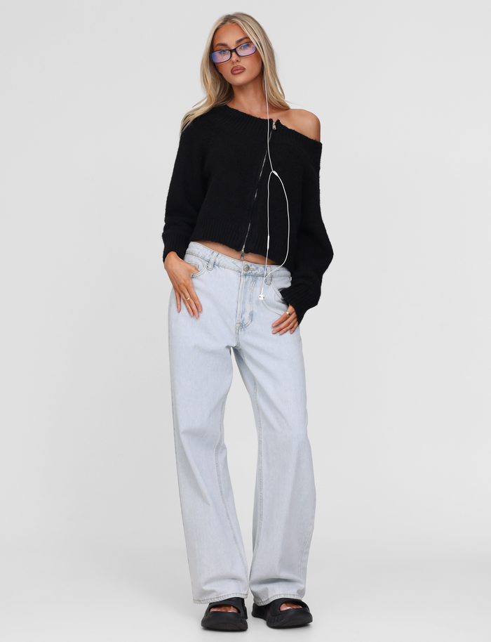 A woman stands against a plain background wearing the TIGERMIST LUNETTE JUMPER in black, paired with light-wash wide-leg jeans, black sandals, glasses, and earphones hanging around her neck.