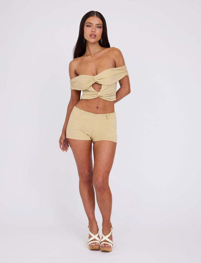 A woman stands against a plain background, wearing the TIGERMIST LYNSEY TOP in gold, an off-shoulder crop top with a front twist and cutout, paired with matching high-waisted shorts. She completes her look with white high-heeled sandals. Her hair is long and straight, and she exudes confidence in her pose.
