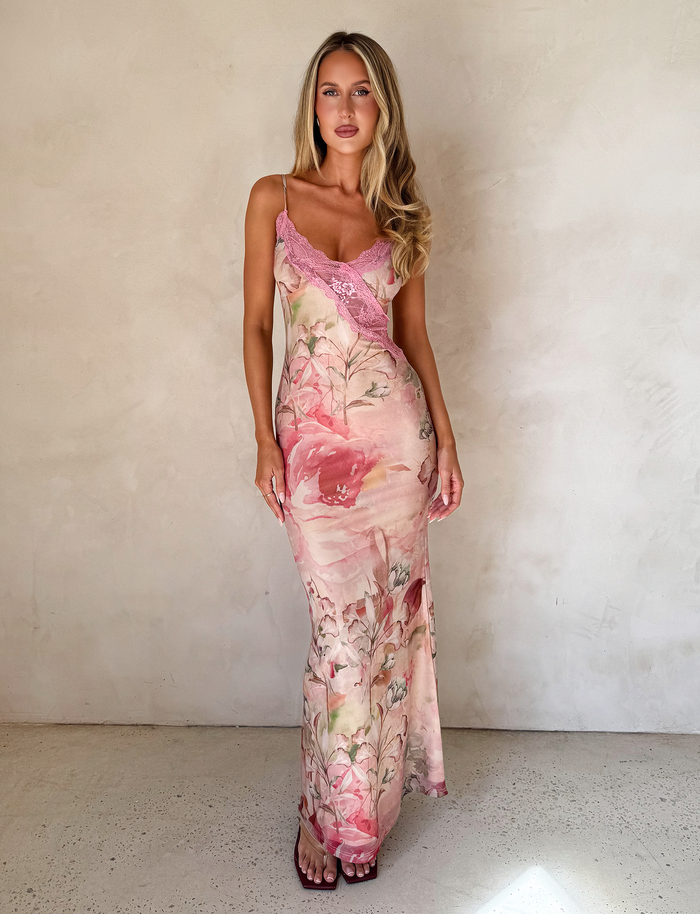 A woman with long blonde hair models the TIGERMIST LYRE MAXI DRESS - WATERCOLOUR FLORAL, featuring adjustable shoulder straps, as she poses against a plain light wall, one hand by her side and the other holding her dress.