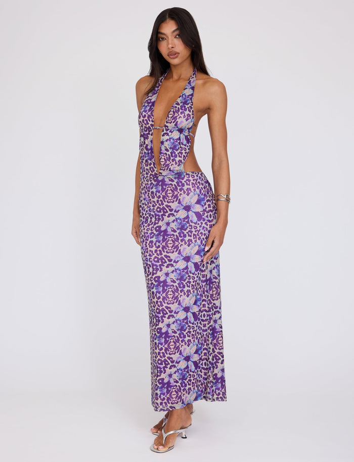 A woman poses in a stunning TIGERMIST MAGDALENA MAXI DRESS, featuring a bold floral pattern by POPPY LEOPARD. The purple, backless halter maxi dress showcases a deep V-neckline and cut-out sides. She is wearing silver, strappy high heels and has a relaxed yet confident expression.