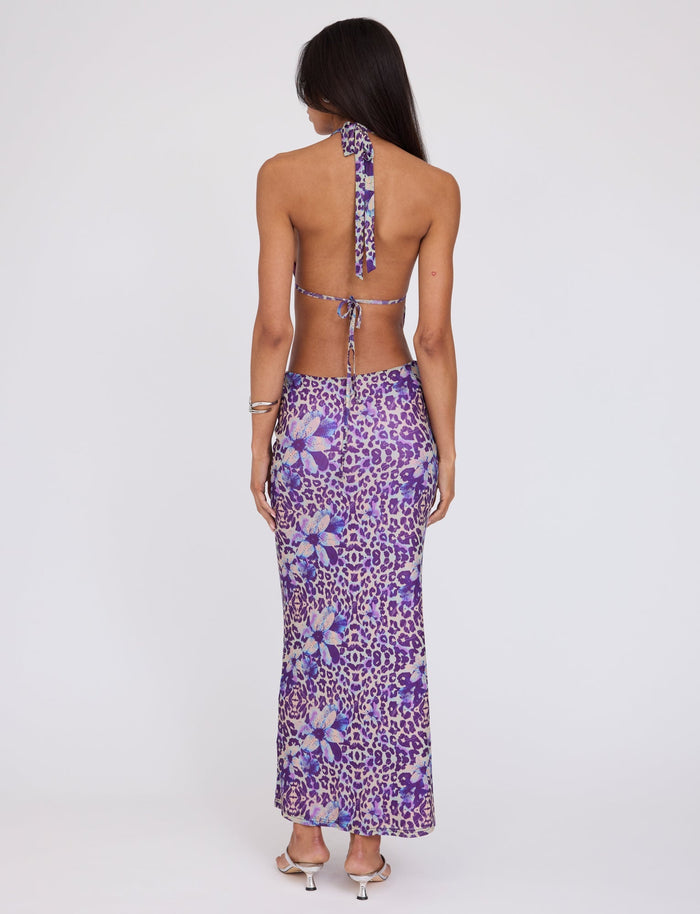 A person with long hair stands with their back to the camera, wearing the Tiger Mist Magdalena Maxi Dress in a purple floral and animal print pattern. The person also dons silver high-heeled sandals and a bracelet on their left wrist.