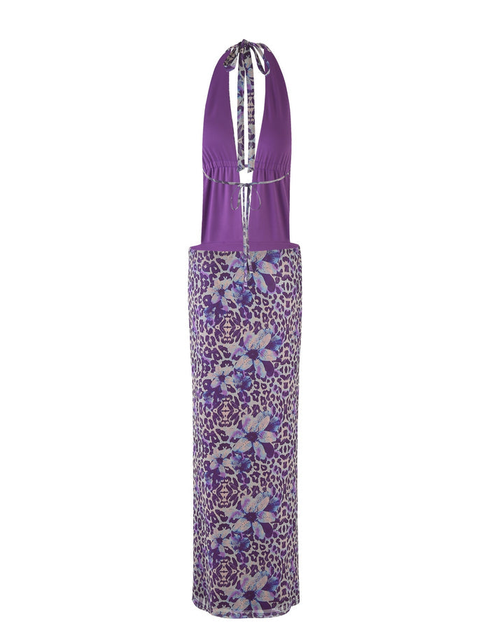 The MAGDALENA MAXI DRESS - PURPLE by TIGERMIST, designed by Poppy Leopard, is a stunning halter-neck dress featuring a plunging neckline and keyhole detail. This dress showcases a fitted bodice and a long, straight skirt embellished with floral and abstract patterns in shades of purple, blue, and white.