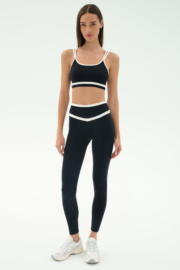 A woman wears the SPLITS59 Mason High Waist Rigor Full Length leggings in Black/White with a matching sports bra and white sneakers, standing against a plain white background.