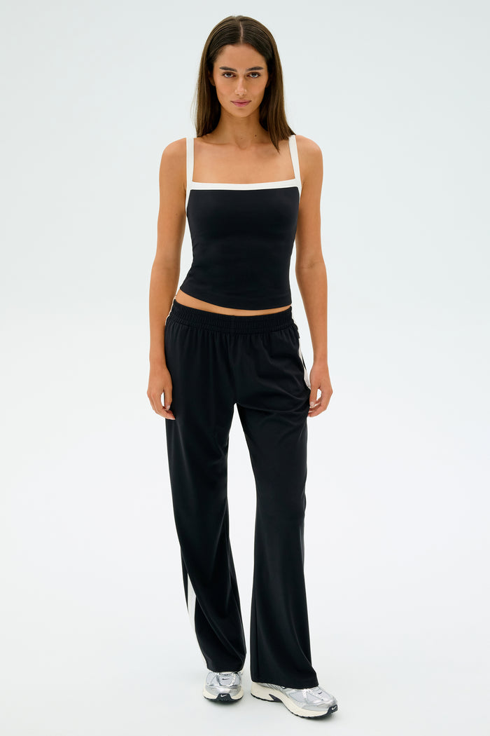A person is standing against a plain background, wearing a black tank top, SPLITS59's Maxie Rigor Track Pant in Black/White, and silver sneakers.