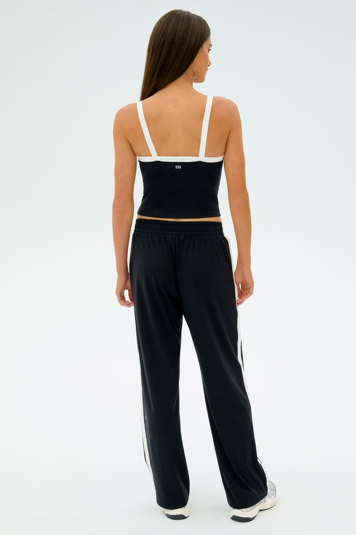 An individual is dressed in a black SPLITS59 sports top featuring white straps, paired with Maxie Rigor Track Pant - Black/White from SPLITS59, standing against a light background.
