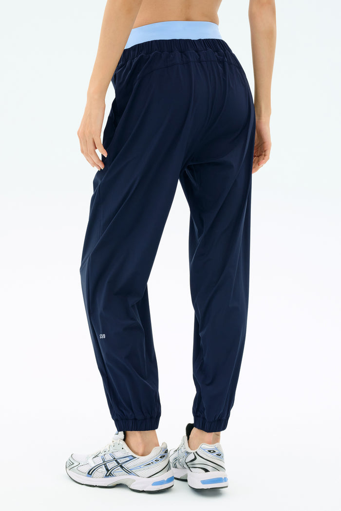 A person wearing SPLITS59 Megan Woven Nylon Jogger in Indigo with a light blue waistband and white athletic shoes, viewed from the back against a plain background.