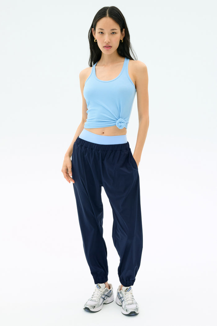A woman wearing a light blue knotted tank top, SPLITS59 Megan Woven Nylon Jogger in Indigo, and white sneakers stands against a plain white background.