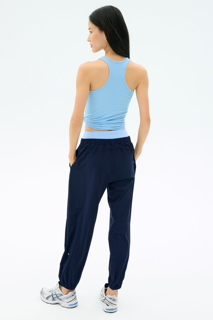 A woman faces away, wearing a light blue sleeveless top, SPLITS59 Megan Woven Nylon Jogger in Indigo, and white sneakers against a plain white background.