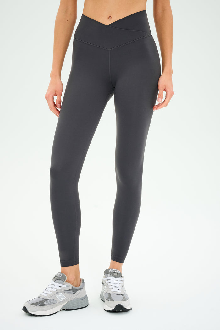 A person wearing SPLITS59′s Mia Rigor 7/8 leggings in Graphite and gray sneakers stands against a plain white background.