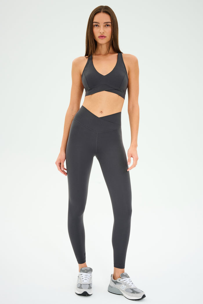 A woman stands against a plain background, dressed in SPLITS59’s Mia Rigor 7/8 Leggings in Graphite, crafted from durable Rigor fabric and paired with a matching dark gray sports bra and gray sneakers.