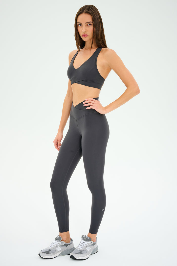 A woman stands in a studio against a plain white background, wearing SPLITS59′s Mia Rigor 7/8 leggings in Graphite crafted from Rigor fabric, paired with a matching dark gray sports bra and silver athletic shoes.