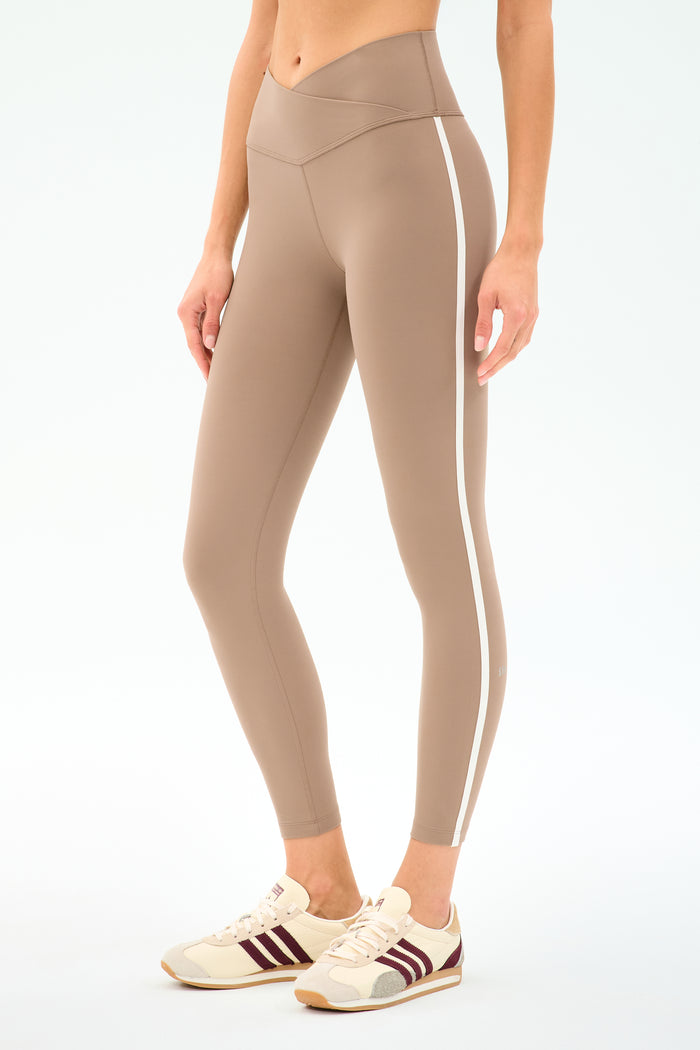 A person wears SPLITS59 Mia Rigor Legging w/ Stripe in Latte/White—high-waisted, stretchy nylon spandex leggings with a white side stripe—paired with cream sneakers featuring maroon stripes, posed against a white background.