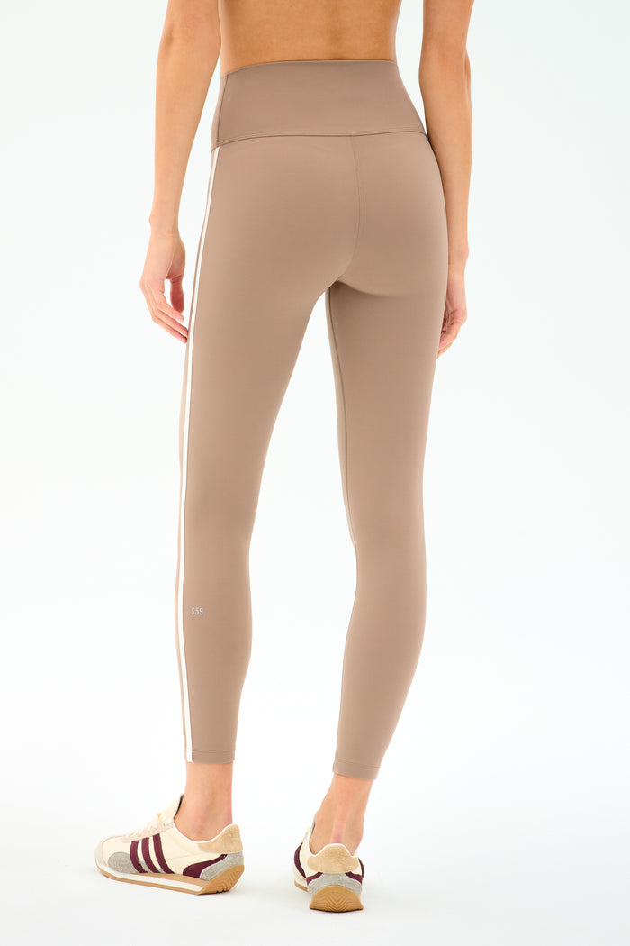 A person wears SPLITS59 Mia Rigor Legging w/ Stripe in Latte/White, high-waisted leggings with a white side stripe, paired with white sneakers with burgundy stripes, standing against a plain white background.