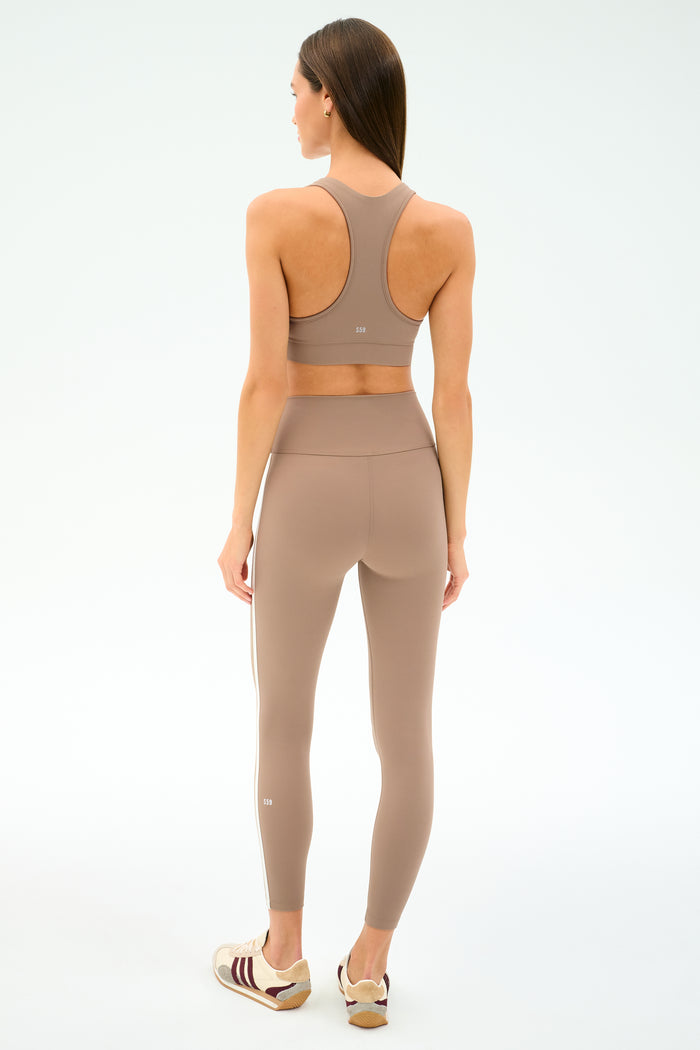 A woman faces away wearing SPLITS59’s Mia Rigor Legging w/ Stripe in Latte/White with a matching sports bra and white-maroon sneakers, set against a plain white background.