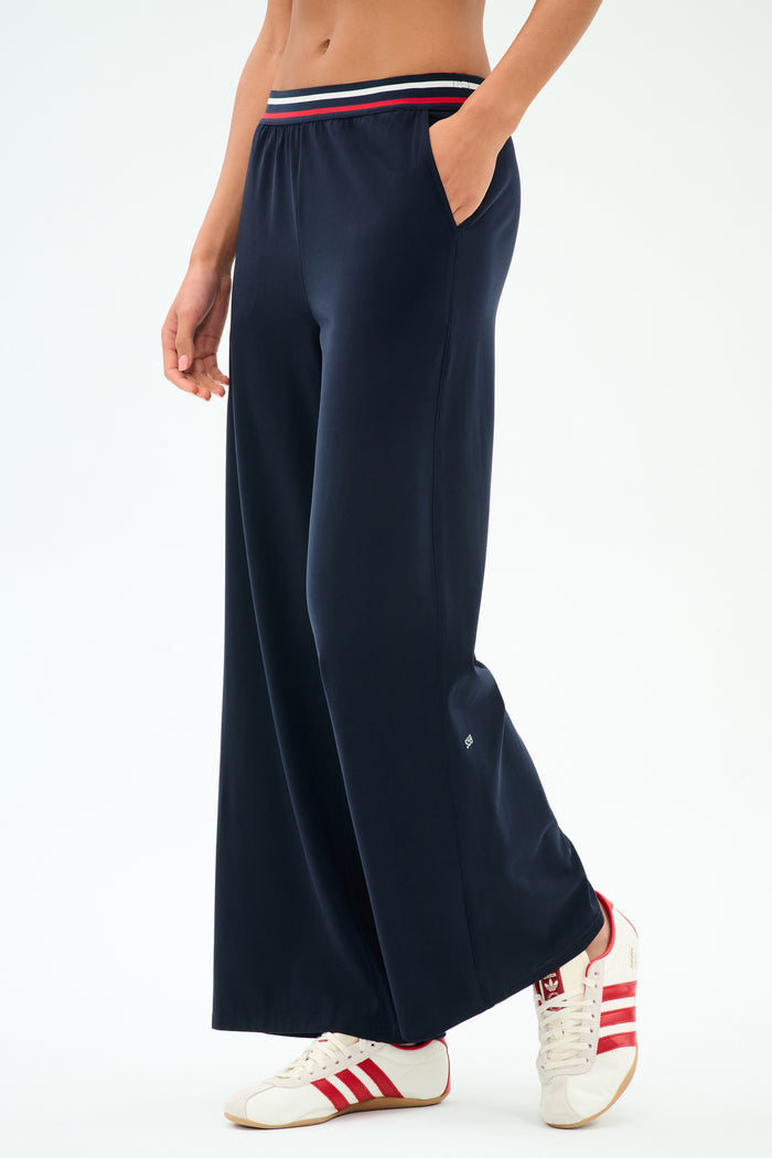 Model is wearing the SPLITS59 Michele Airweight Trouser in Indigo with a striped waistband and white sneakers featuring red stripes, shown from shoulders down with hands in pockets.